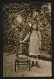 Photographie d'une jeune fille, debout à côté d'une chaise vide, dans le jardin, non datée