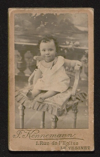 Photographie d'un bébé, en robe, assis sur une chaise, non datée